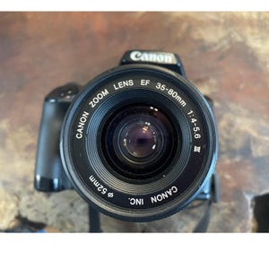 May include: Black Canon camera with a zoom lens. The lens is marked "Canon Zoom Lens EF 35-80mm 1:4-5.6 III" and has a 52mm diameter. The camera body is black with the Canon logo on top.