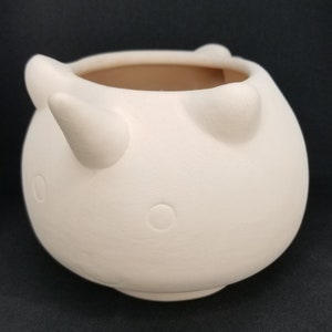 Puede incluir: Maceta de cerámica blanca con forma de toro con dos cuernos y dos pequeños círculos para los ojos.