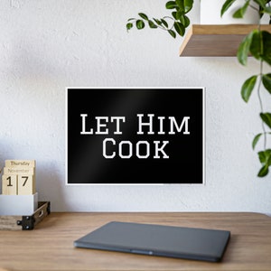 Peut inclure: Une affiche noire et blanche avec le texte "LET HIM COOK" en gras, dans une police de style universitaire. L'affiche est encadrée en blanc et exposée sur un bureau en bois. Un calendrier et un ordinateur portable sont également visibles.