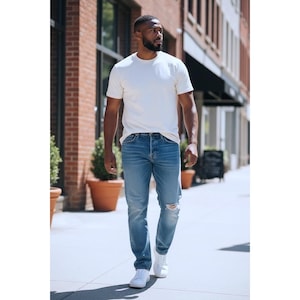 Könnte beinhalten: Eine Person trägt ein cremefarbenes Rundhals-T-Shirt, blaue Jeans mit einem Knie-Riss und weiße Turnschuhe. Die Person geht auf einem Bürgersteig vor einem Backsteingebäude.