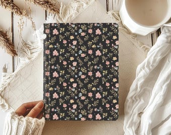 May include: A black journal with a floral pattern featuring pink, yellow, and blue flowers. The journal is held by a person wearing a white sweater. A white mug and a beige cloth are also in the image.