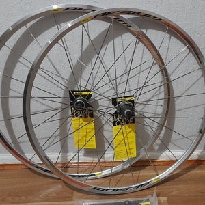 May include: A pair of silver bicycle wheels with black spokes and black rim tape. The wheels are labelled "Aksium" and have yellow stickers with the text "Mavic Care".