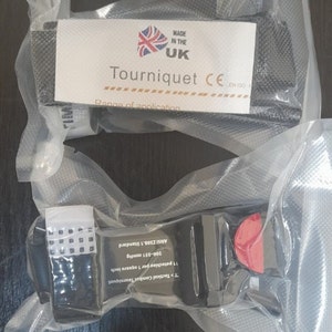 Könnte beinhalten: Ein schwarzer taktischer Kampftourniquet mit einer roten Entriegelungsschnalle. Der Tourniquet ist mit "T>Tactical Combat Tourniquet", "11 petechiae per 1 square inch", "200-351 mmHg" und "ANSI Z308.1 Standard" beschriftet.