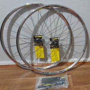 May include: A pair of silver bicycle wheels with black rims and spokes. The wheels are stacked on top of each other and are sitting on a wooden floor. There are two yellow and black labels that say "Mavic Care" on them. There is a clear plastic bag with black and silver hardware in the bottom right corner of the image.