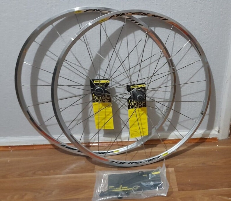 May include: A pair of silver bicycle wheels with black rims and spokes. The wheels are stacked on top of each other and are sitting on a wooden floor. There are two yellow and black labels that say "Mavic Care" on them. There is a clear plastic bag with black and silver hardware in the bottom right corner of the image.