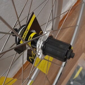 May include: Close-up of a silver bicycle wheel with black spokes and a black hub. The hub has a black Mavic logo on it.