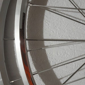May include: A silver bicycle wheel with black and orange accents. The wheel has a deep rim and is made of metal. The spokes are thin and silver. The wheel is mounted on a white wall.