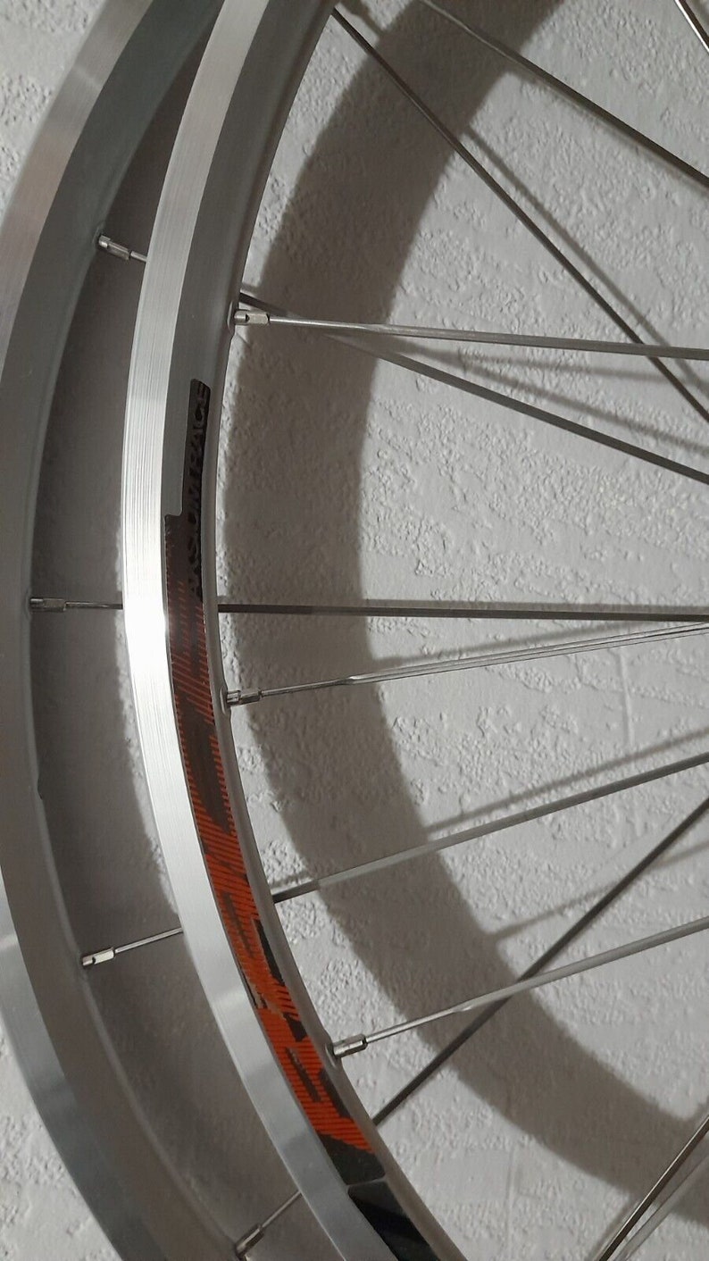 May include: A silver bicycle wheel with black and orange accents. The wheel has a deep rim and is made of metal. The spokes are thin and silver. The wheel is mounted on a white wall.