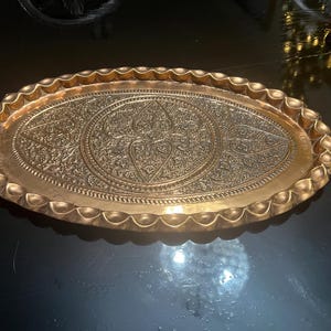 Puede incluir: Una bandeja ovalada de metal dorado con intrincados patrones florales y geométricos en relieve. La bandeja tiene un borde festoneado y un acabado pulido, adecuada para servir o exhibir.