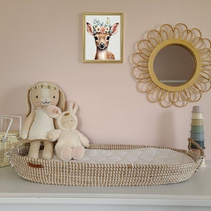Pu&ograve; includere: Un lettino in vimini con una fodera imbottita bianca, un coniglio di peluche bianco e un coniglio di peluche marrone con un cerchietto floreale. Il lettino &egrave; su una superficie bianca con un cesto in vimini, una pila di bicchieri impilabili in silicone colorati e uno specchio rotondo in rattan sul muro dietro.