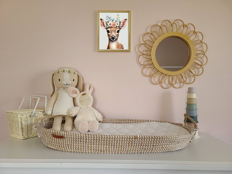 Pu&ograve; includere: Un lettino in vimini con una fodera imbottita bianca, un coniglio di peluche bianco e un coniglio di peluche marrone con un cerchietto floreale. Il lettino &egrave; su una superficie bianca con un cesto in vimini, una pila di bicchieri impilabili in silicone colorati e uno specchio rotondo in rattan sul muro dietro.