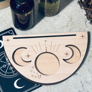 May include: A wooden crescent moon incense holder with three compartments, two for incense sticks and one for a larger incense cone. The holder is decorated with a moon, stars, and a sun design.