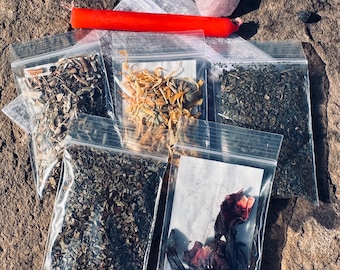 May include: Five small clear plastic bags filled with dried herbs and flowers. The bags are arranged on a brown, rocky surface. A red candle and a pink crystal are also visible in the image.