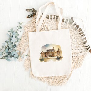 Peut inclure: Un sac fourre-tout en toile blanche avec une illustration aquarelle d'un grand b&acirc;timent &agrave; fa&ccedil;ade dor&eacute;e. Le b&acirc;timent est situ&eacute; sur un fond de ciel bleu clair avec quelques nuages. Le sac fourre-tout a une texture de tissu naturelle.