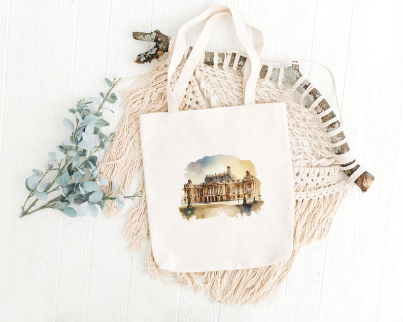 Peut inclure: Un sac fourre-tout en toile blanche avec une illustration aquarelle d'un grand b&acirc;timent &agrave; fa&ccedil;ade dor&eacute;e. Le b&acirc;timent est situ&eacute; sur un fond de ciel bleu clair avec quelques nuages. Le sac fourre-tout a une texture de tissu naturelle.