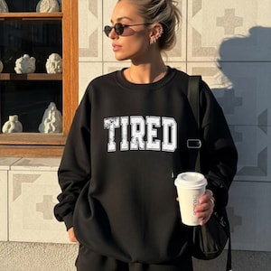 May include: Black sweatshirt with the word "TIRED" in distressed white lettering. The person is wearing black shorts, sunglasses, and has a black bag. They are holding a white coffee cup.