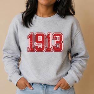 May include: A light grey crewneck sweatshirt featuring the number 1913 printed in a large, bold, red varsity-style font with a distressed, weathered texture. The garment has long sleeves with ribbed cuffs and a ribbed neckline.