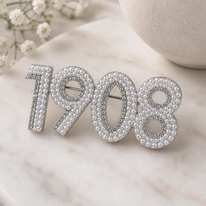 May include: A silver-toned brooch in the shape of the numbers "1908". The numbers are encrusted with small, white faux pearls. The "1" and the "8" are also accented with clear rhinestones. The brooch is set against a marble surface.