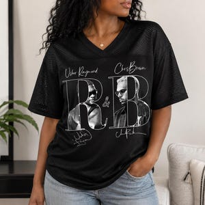 May include: A black mesh V-neck t-shirt featuring a graphic print of Usher Raymond and Chris Brown. The design includes the text "Usher Raymond" and "Chris Brown" above their portraits, a large "R&B" in the centre, and their signatures below.