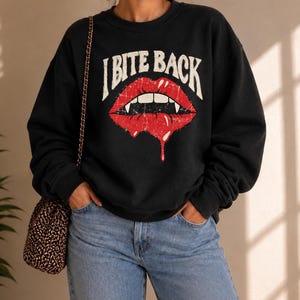Könnte beinhalten: Ein schwarzes Langarm-Sweatshirt mit einem Grafikdruck von roten Lippen mit weißen Fangzähnen und dem Text I BITE BACK in einer abgenutzten weißen Schriftart. Das Design zeigt einen tropfenden roten Flüssigkeitseffekt auf der Unterlippe.