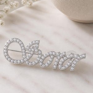 May include: A silver-toned brooch spelling out the word "lover" in cursive script. The letters are outlined with small, round, white pearls. The brooch has a pin backing for secure attachment.