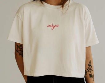 Pode incluir: Uma t-shirt de manga curta na cor creme com a palavra virgo escrita numa fonte cursiva vermelha centrada no peito. A peça tem um decote redondo e um corte curto e quadrado, proporcionando uma estética simples e limpa.