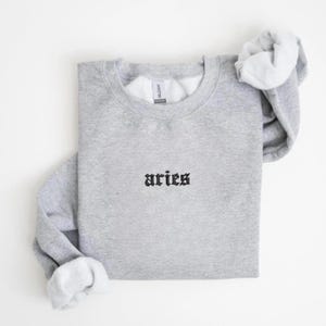 May include: A light gray sweatshirt with the word "aries" embroidered in black gothic font. The sweatshirt has a crew neck and long sleeves, with the sleeves and neck rolled up. The inside of the sleeves and neck are white.