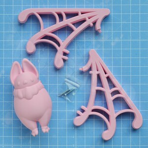 A flat lay showcasing all of the pieces that come with purchase. One stylized sculptural round bat body piece, two spiderweb/cobweb shaped wing pieces, and a small plastic baggie containing two #4 phillips head wood screws, on top of a light blue gridded background