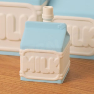 May include: A light blue and white milk carton-shaped bottle stopper. The word "MILK" is prominently displayed on the front. The stopper has a small, cylindrical top. The background features a similar design.