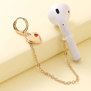May include: A gold chain with a heart charm attached to a white wireless earbud. The chain is connected to a hoop earring.