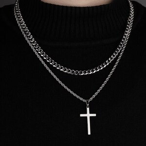 Puede incluir: Un collar de cadena de plata con un colgante de cruz de plata. El collar está formado por dos cadenas, una más gruesa que la otra.