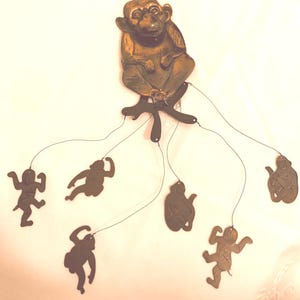 May include: A bronze-colored monkey mobile with six smaller monkey figures suspended by strings. The main monkey figure is seated, with its hand to its face. The smaller monkeys are in various poses, creating a playful and decorative piece.