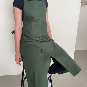 May include: A dark green apron with a large front pocket and cross-back straps. The apron is knee-length and has a split design on the front. The person is wearing a black t-shirt. The apron is made of a smooth, solid-coloured fabric.