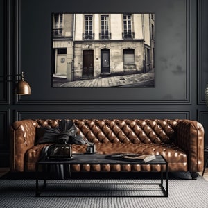 May include: A brown leather sofa with tufted upholstery sits in a dark room with black walls and a black and white photograph of a Parisian street scene hanging above it.