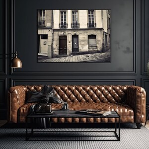 May include: A brown leather sofa with tufted upholstery sits in a dark room with black walls and a black and white photo of a Parisian street hanging on the wall.