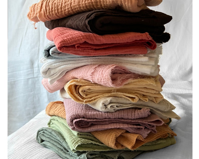 May include: A stack of 12 different colored fabric squares, each with a slightly different texture. The fabrics are arranged in a pile, with the top fabric being a light brown color. The fabrics are all soft and appear to be made of a natural material.