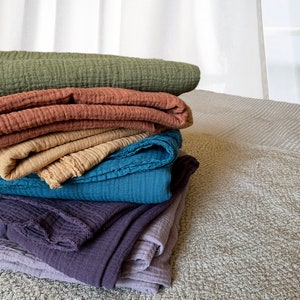 May include: A stack of five soft, textured blankets in various shades of green, brown, beige, blue, and purple. The blankets are folded and arranged on a light gray textured surface.