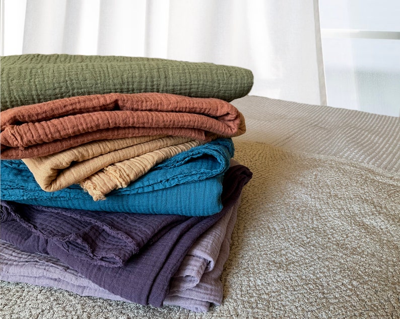 May include: A stack of five soft, textured blankets in various shades of green, brown, beige, blue, and purple. The blankets are folded and arranged on a light gray textured surface.