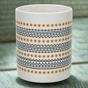 May include: White ceramic mug with a black and yellow geometric pattern. The pattern features rows of small yellow flowers, wavy lines, and straight lines.