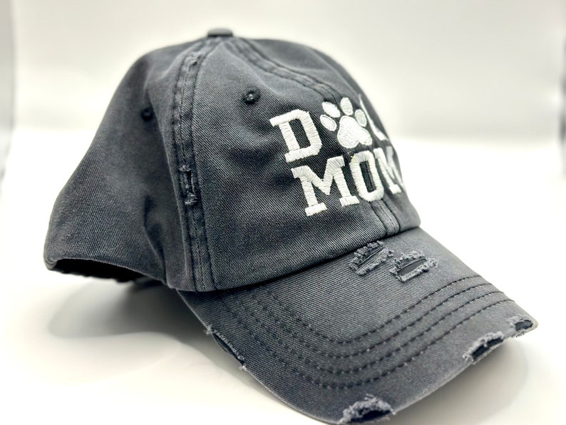 K&ouml;nnte beinhalten: Eine graue, abgenutzte Baseballkappe mit wei&szlig;er Stickerei, auf der "Dog Mom" mit einem Pfotenabdruck steht.