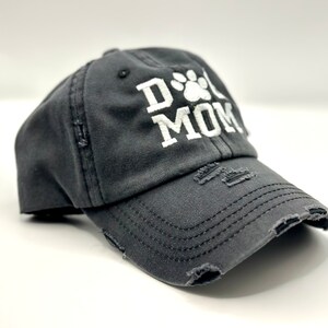 K&ouml;nnte beinhalten: Eine schwarze, abgenutzte Baseballkappe mit einem wei&szlig;en gestickten Pfotenabdruck und dem Text "Dog Mom".