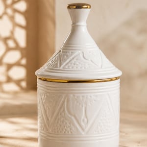 Op de afbeelding: Een witte keramische pot met een bijpassend conisch deksel, voorzien van reliëf geometrische patronen en getextureerde driehoekige details. Het deksel is bekroond met een kleine goudkleurige knop en een dunne gouden band siert de rand.