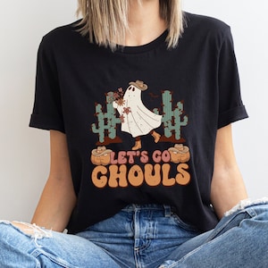 May include: Black t-shirt with a ghost wearing a cowboy hat and holding flowers. The ghost is standing in front of cacti and pumpkins. The text "Let's Go Ghouls" is printed on the shirt.