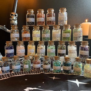 Puede incluir: Una colección de pequeños frascos de vidrio llenos de varias piedras preciosas, cada uno etiquetado. Los frascos están dispuestos sobre una superficie negra, con velas y un soporte decorativo de calavera en el fondo. Las piedras preciosas incluyen amatista, cuarzo y ópalo.