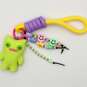 May include: A quirky keychain featuring a lime green monster charm, a purple and yellow rope loop, and a beaded strand with smiley faces and flower beads. The monster has a red mouth and sleepy eyes.
