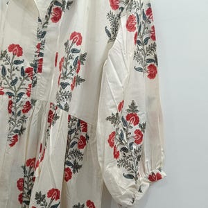 May include: A white cotton dress with a floral print in shades of red and green. The dress has a V-neckline and long sleeves with a gathered detail at the shoulders.