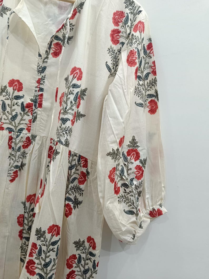 May include: A white cotton dress with a floral print in shades of red and green. The dress has a V-neckline and long sleeves with a gathered detail at the shoulders.