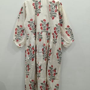 May include: A white cotton dress with a floral print featuring red and green flowers. The dress has a V-neck and long sleeves.