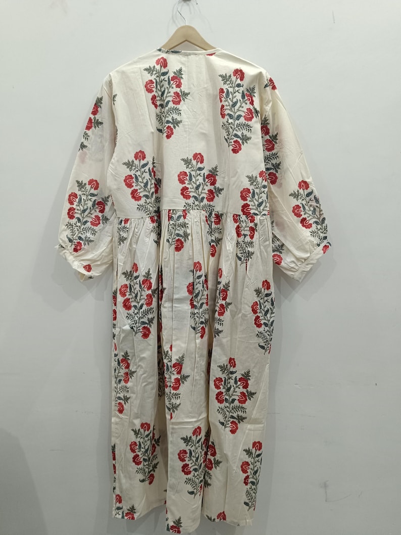 May include: A white cotton dress with a floral print featuring red and green flowers. The dress has a V-neck and long sleeves.