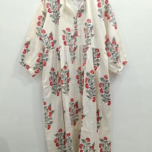 May include: A white cotton dress with a floral print in shades of red and green. The dress has a V-neckline and a tie at the neckline. The sleeves are three-quarter length and the dress has a gathered skirt.